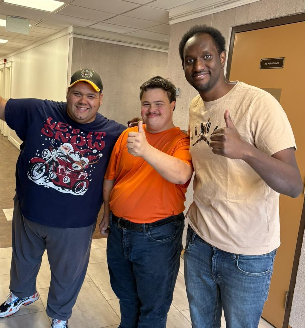 Three people from Adults in Motion Hamilton smiling together in a hallway, two of them giving a thumbs-up.