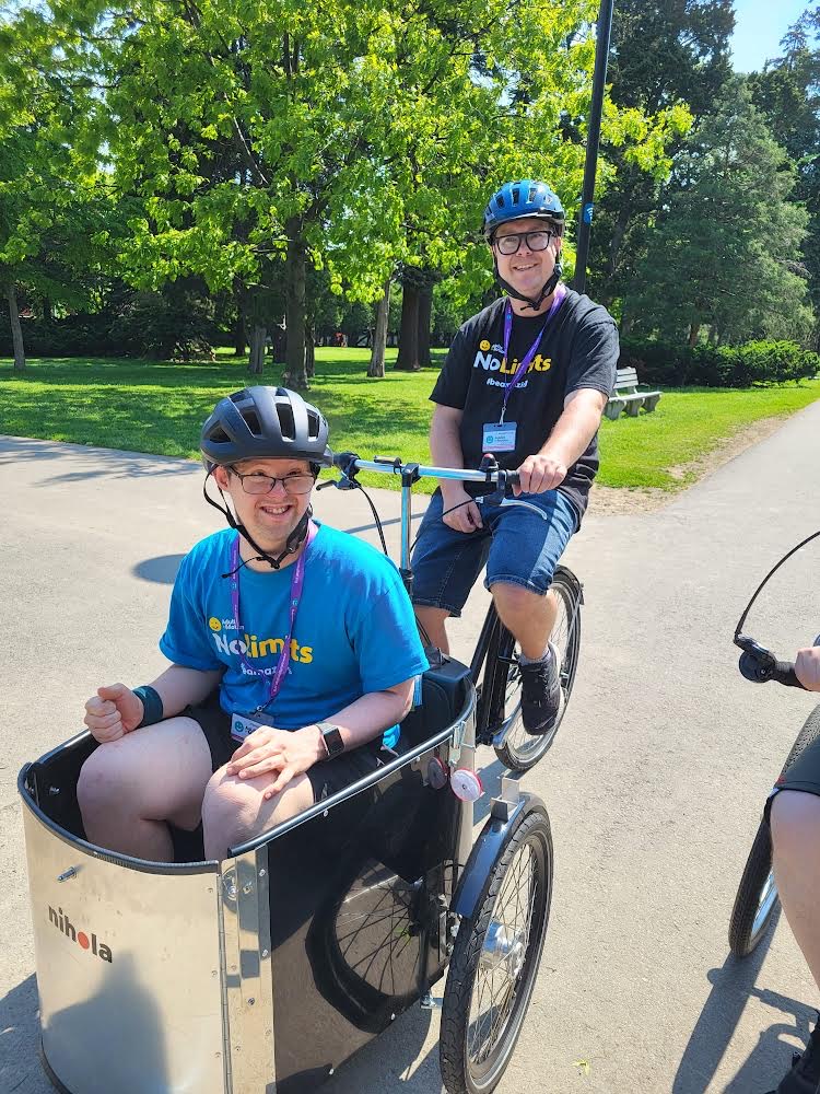 Two members ride adaptive bikes through a park with the Oakville day program.